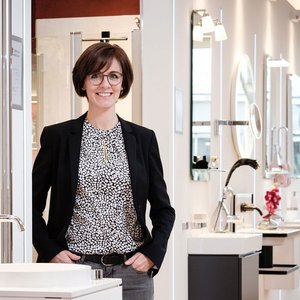 In einem stilvollen Ausstellungsraum präsentiert eine Frau in einem Blazer Badezimmeraccessoires und lächelt freundlich in die Kamera.