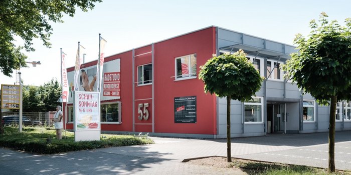 Ein modernes Gebäude mit roter Fassade und großen Fenstern, umgeben von Bäumen und einem gepflegten Parkplatz.