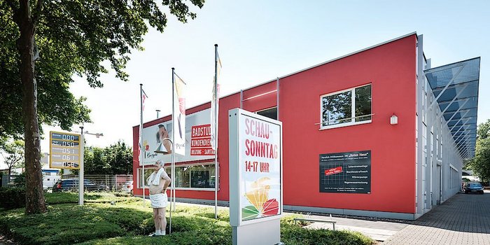 Ein modernes, rotes Gebäude mit großen Fenstern und Werbeschildern, umgeben von Bäumen und einem klaren Himmel.