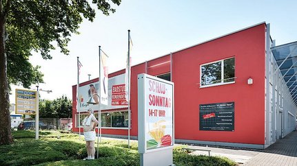 Moderne Architektur eines Sanitärgeschäfts in der Stadt Ein modernes, rotes Gebäude mit großen Fenstern und Werbeschildern, umgeben von Bäumen und einem klaren Himmel.