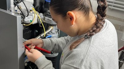 In einem technischen Raum repariert ein Mädchen mit Zopf eine Maschine, während sie mit einer Zange Kabel bearbeitet.