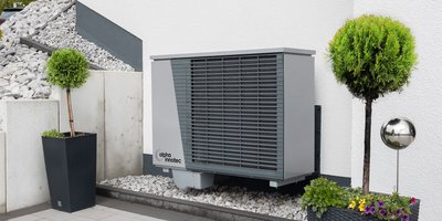Ein graues Klimagerät steht neben einem modernen Pflanzgefäß mit einem runden, grünen Baum und bunten Blumen.