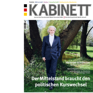 Ein älterer Mann mit grauen Haaren steht in einem Park, umgeben von Bäumen und grünem Gras, in eleganter Kleidung.