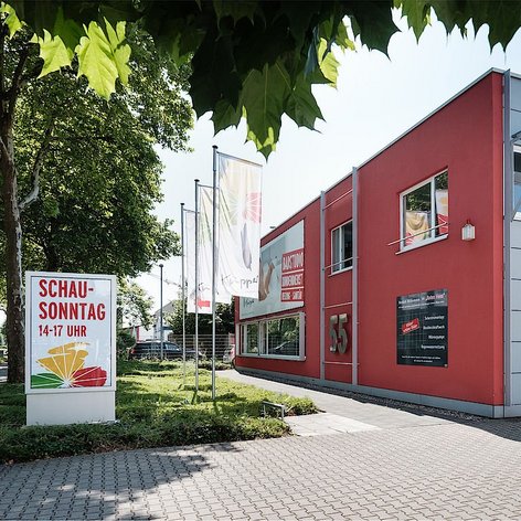 Ein rotes Gebäude mit großen Fenstern und einem Schild, das einen Schautag von 14 bis 17 Uhr ankündigt.