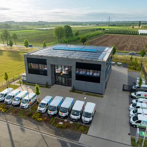 Ein modernes Bürogebäude mit Solarpanelen auf dem Dach, umgeben von grünen Feldern und einer Reihe von Lieferwagen.