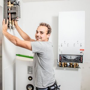 Ein Handwerker installiert eine Heizungsanlage, während er lächelt und mit einer Hand ein Gerät hält.