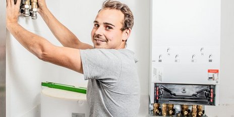 Ein Handwerker installiert eine Heizungsanlage, während er lächelt und mit einer Hand ein Gerät hält.