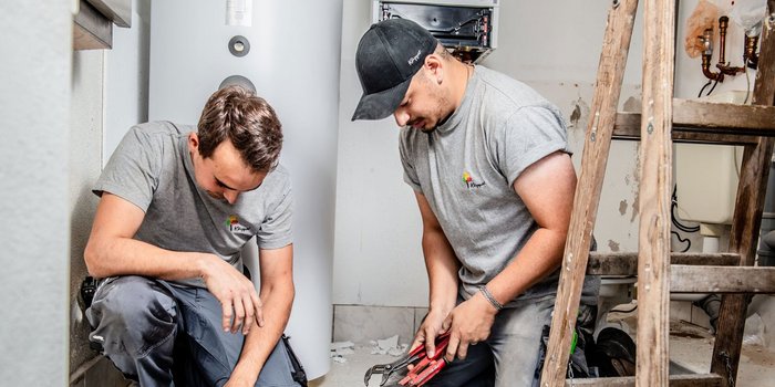 Ein Handwerker kniet und überprüft die Rohre, während sein Kollege mit einer Zange an einem anderen Teil arbeitet.