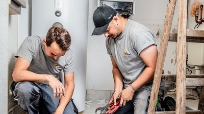Ein Handwerker kniet und überprüft die Rohre, während sein Kollege mit einer Zange an einem anderen Teil arbeitet.