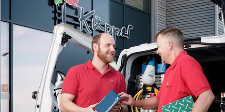 Zwei Männer in roten Hemden stehen an einem Lieferwagen und lächeln sich an, während sie ein Paket austauschen.