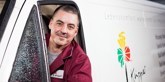 Ein lächelnder Mann mit Schnurrbart schaut aus dem Fenster eines Lieferwagens, während Regentropfen die Scheibe benetzen.