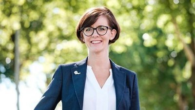 Eine lächelnde Frau mit Brille steht vor einem grünen Hintergrund aus Bäumen, gekleidet in einem eleganten Blazer.