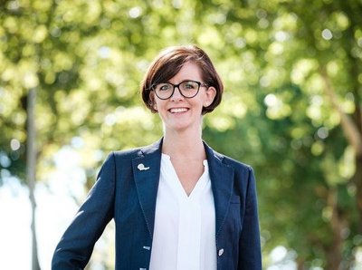 Eine lächelnde Frau mit Brille steht vor einem grünen Hintergrund aus Bäumen, gekleidet in einem eleganten Blazer.