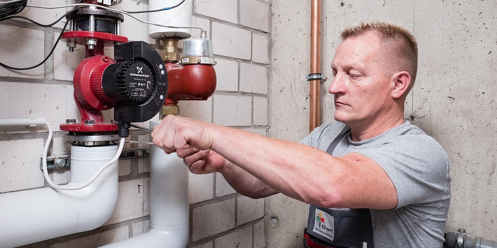 Ein Handwerker arbeitet konzentriert an einer roten Pumpe, während er ein Werkzeug in der Hand hält und an Rohren schraubt.