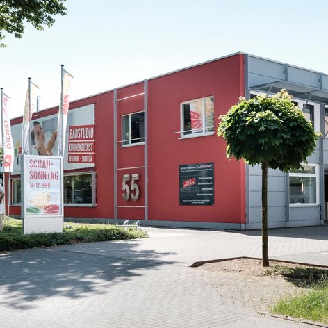Ein modernes, rotes Gebäude mit großen Fenstern und der Nummer 55, umgeben von Bäumen und einem gepflasterten Weg.