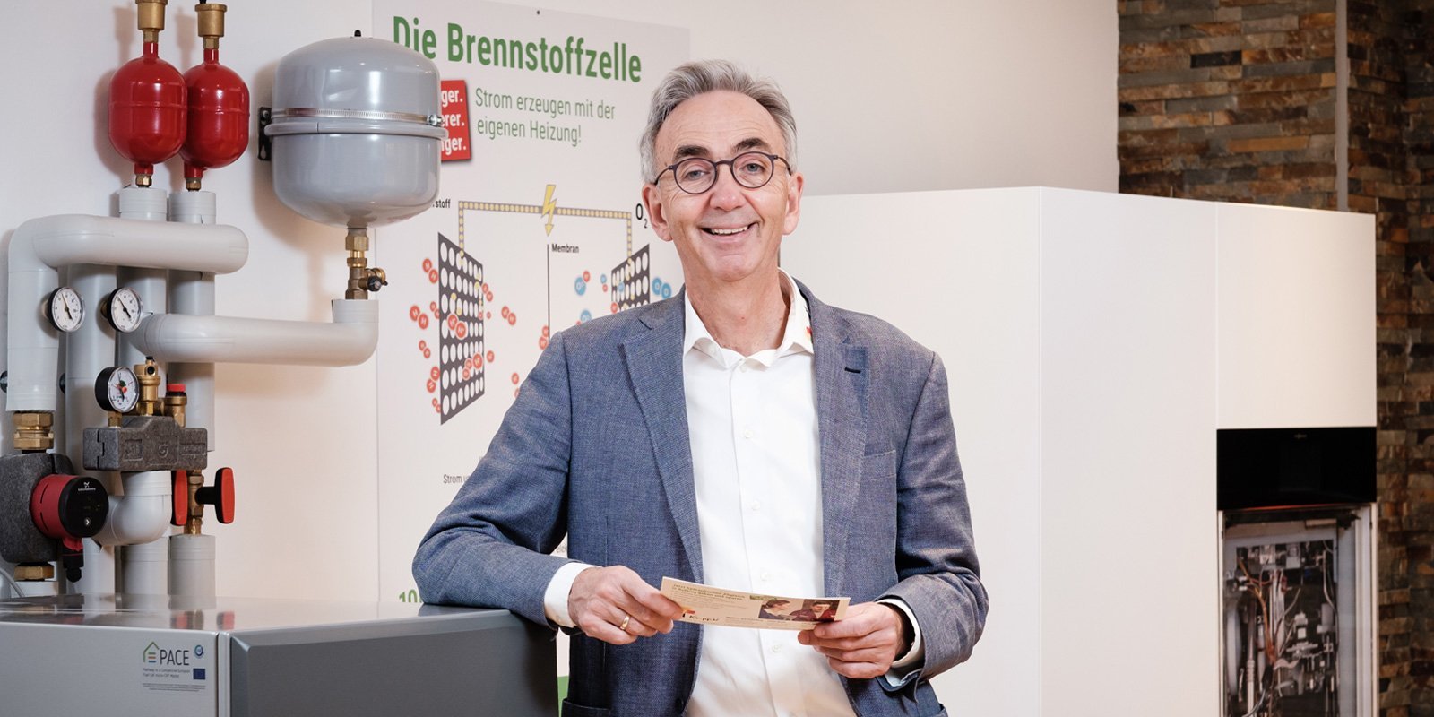 Ein lächelnder Mann steht neben einer modernen Heizungsanlage und hält ein Informationsblatt in der Hand.