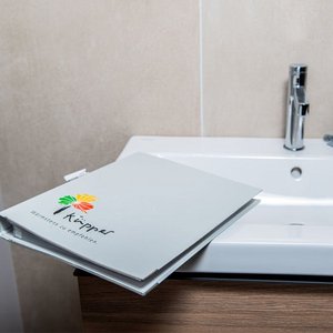 Der graue Ordner mit dem Küpper-Logo ist auf einem stilvollen Waschbecken platziert, umgeben von neutralen Wandfliesen.