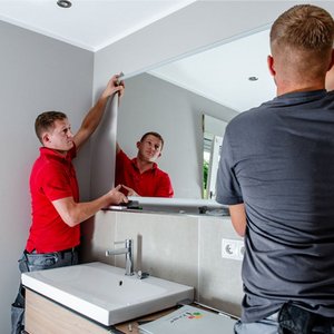 Ein Handwerker in einem roten Shirt hält einen Spiegel, während sein Kollege ihn an der Wand anbringt, umgeben von Tageslicht.