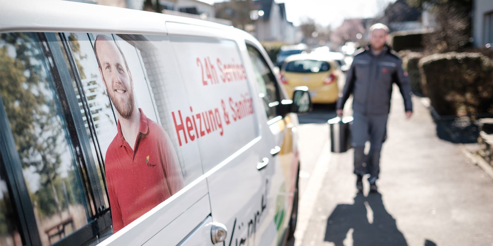 Ein Handwerker in grauer Arbeitskleidung trägt einen Eimer und geht auf einem Bürgersteig entlang, um einen Service zu leisten.