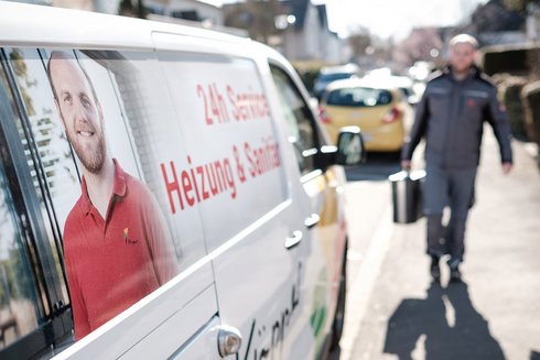 Ein Handwerker in grauer Arbeitskleidung trägt einen Eimer und geht auf einem Bürgersteig entlang, um einen Service zu leisten.