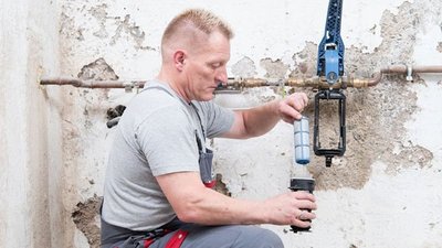 Ein Handwerker installiert einen Wasserfilter an einer Wand mit sichtbaren Schäden und Rohren im Hintergrund.