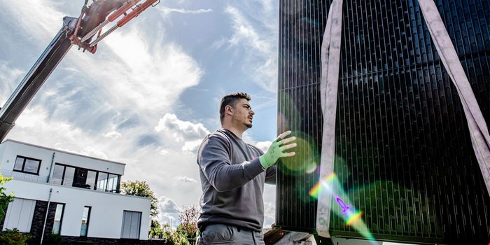 Ein Arbeiter mit grünen Handschuhen positioniert ein großes, schwarzes Objekt unter einem blauen Himmel mit Wolken.