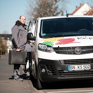 Der Fahrer in grauer Arbeitskleidung lächelt, während er an einem bunten Lieferwagen steht, der auf einer ruhigen Straße parkt.