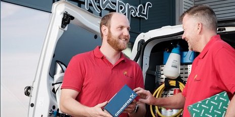 Zwei Männer in roten Shirts stehen an einem Lieferwagen und tauschen freundlich eine Kiste aus, während sie lächeln.