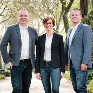Drei Personen stehen auf einem Weg, umgeben von Bäumen, lächeln freundlich in die Kamera und tragen formelle Kleidung.