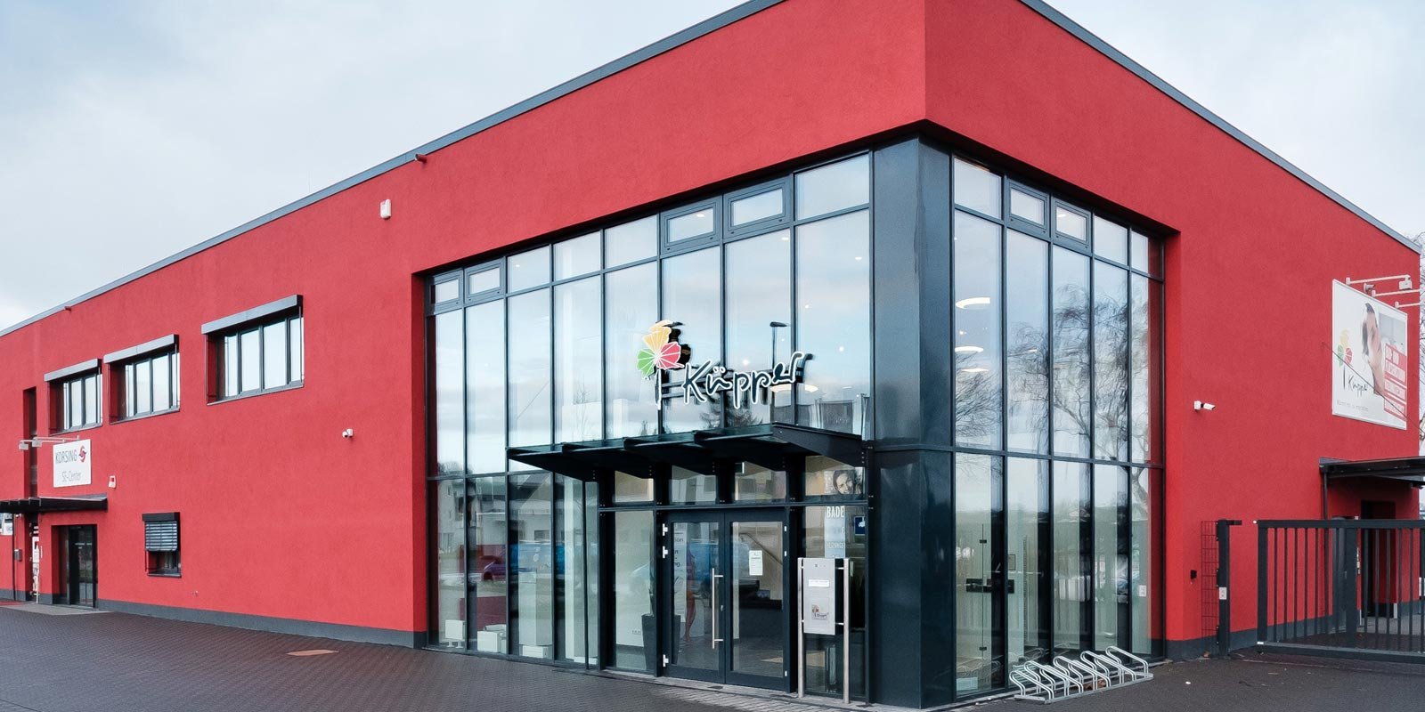 Ein modernes Gebäude mit roter Fassade und großen Fenstern, das einladend wirkt und einen klaren Zugang bietet.