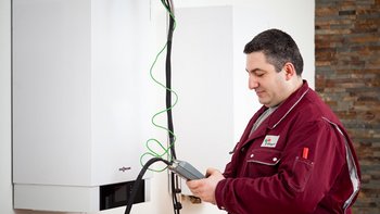 Ein Techniker in einem roten Arbeitsanzug überprüft ein Heizgerät mit einem Messgerät in der Hand.