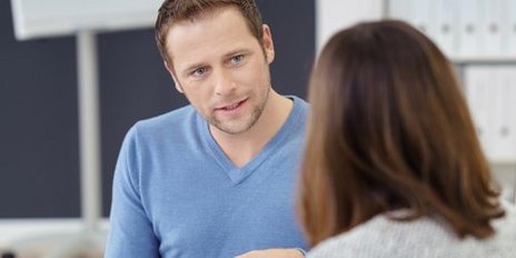 Ein Mann in einem blauen Pullover spricht engagiert mit einer Frau, während sie in einem modernen Büro sitzen.