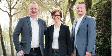 Drei Personen stehen lächelnd im Freien, umgeben von Bäumen, und tragen formelle Kleidung, die Professionalität ausstrahlt.