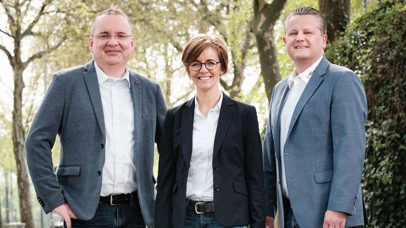 Drei Personen stehen lächelnd im Freien, umgeben von Bäumen, und tragen formelle Kleidung, die Professionalität ausstrahlt.