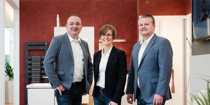 Drei Personen in formeller Kleidung stehen in einem modernen Raum mit roter Wand und stilvollen Möbeln.