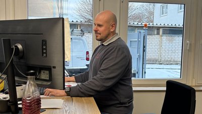 Ein Mann sitzt an einem Schreibtisch und arbeitet konzentriert an einem Computer, während er durch ein Fenster nach draußen schaut.