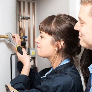 Eine junge Frau arbeitet konzentriert mit einem Schraubenschlüssel an einer Heizungsinstallation, während ein Mann zuschaut.